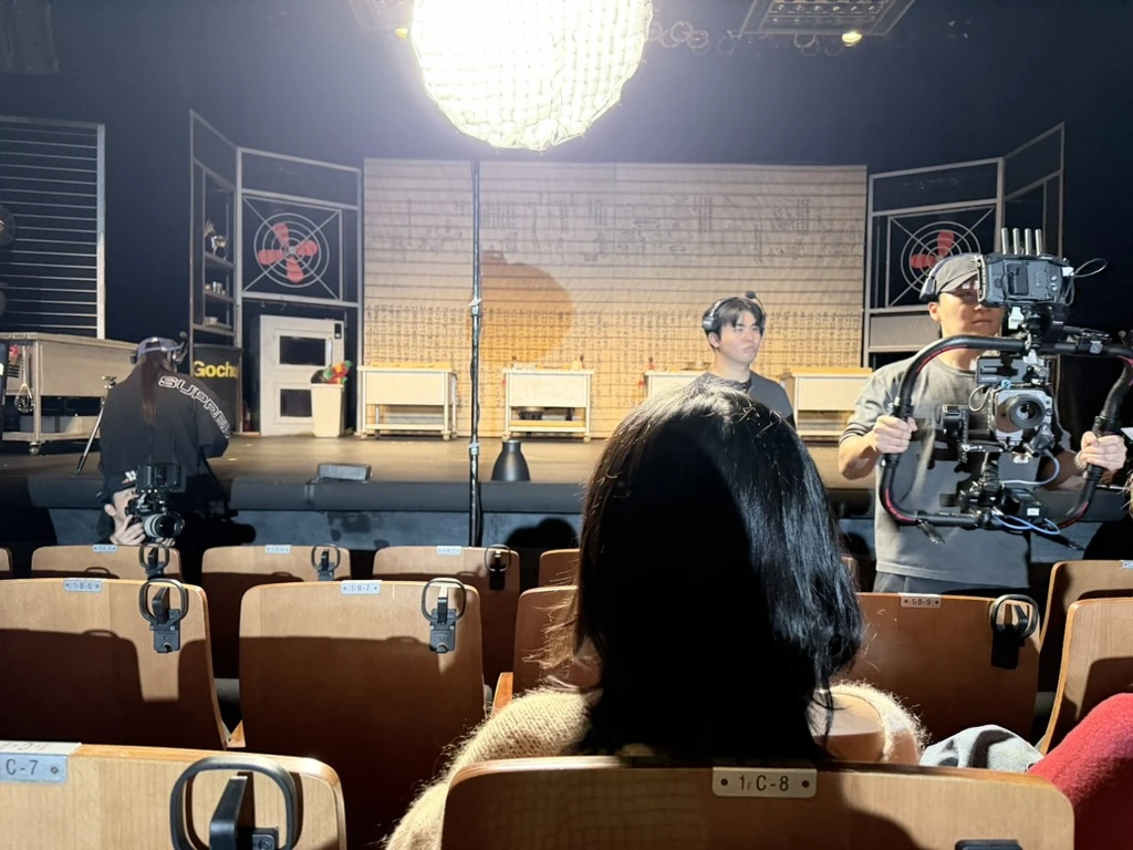 A person seated in a theater facing a production crew and camera equipment near the stage