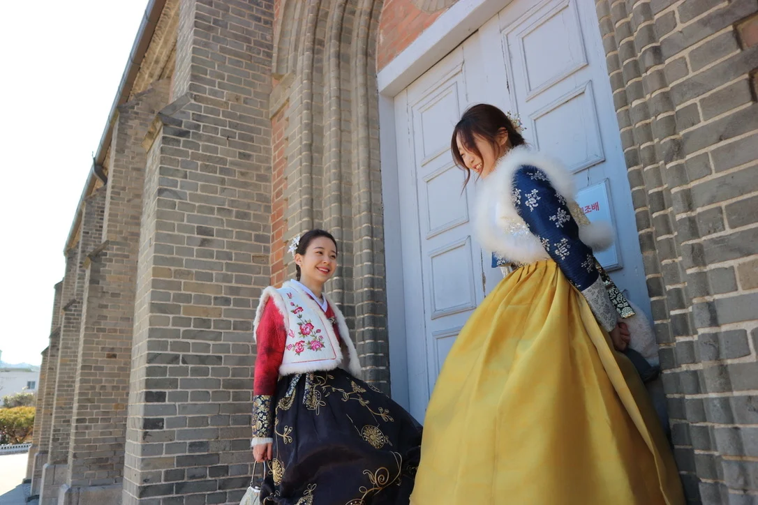 Two models in embroidered winter hanbok beside a brick building