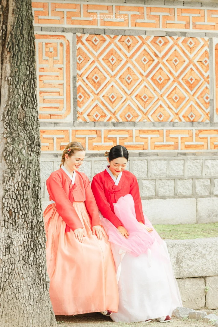 Two models in hanbok seated beside a patterned wall